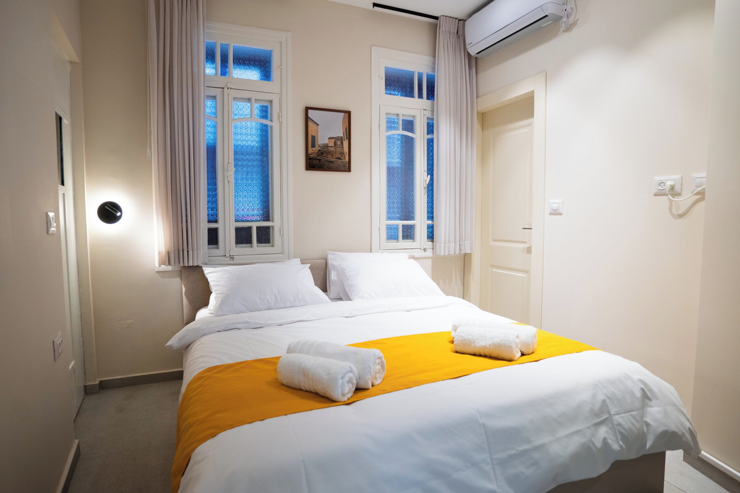 Habitación de hotel con cama, sábanas blancas, una manta amarilla y ventana con vista a un edificio.