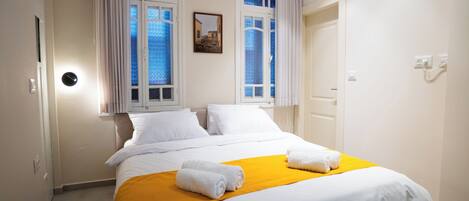 Habitación de hotel con cama, sábanas blancas, una manta amarilla y ventana con vista a un edificio.