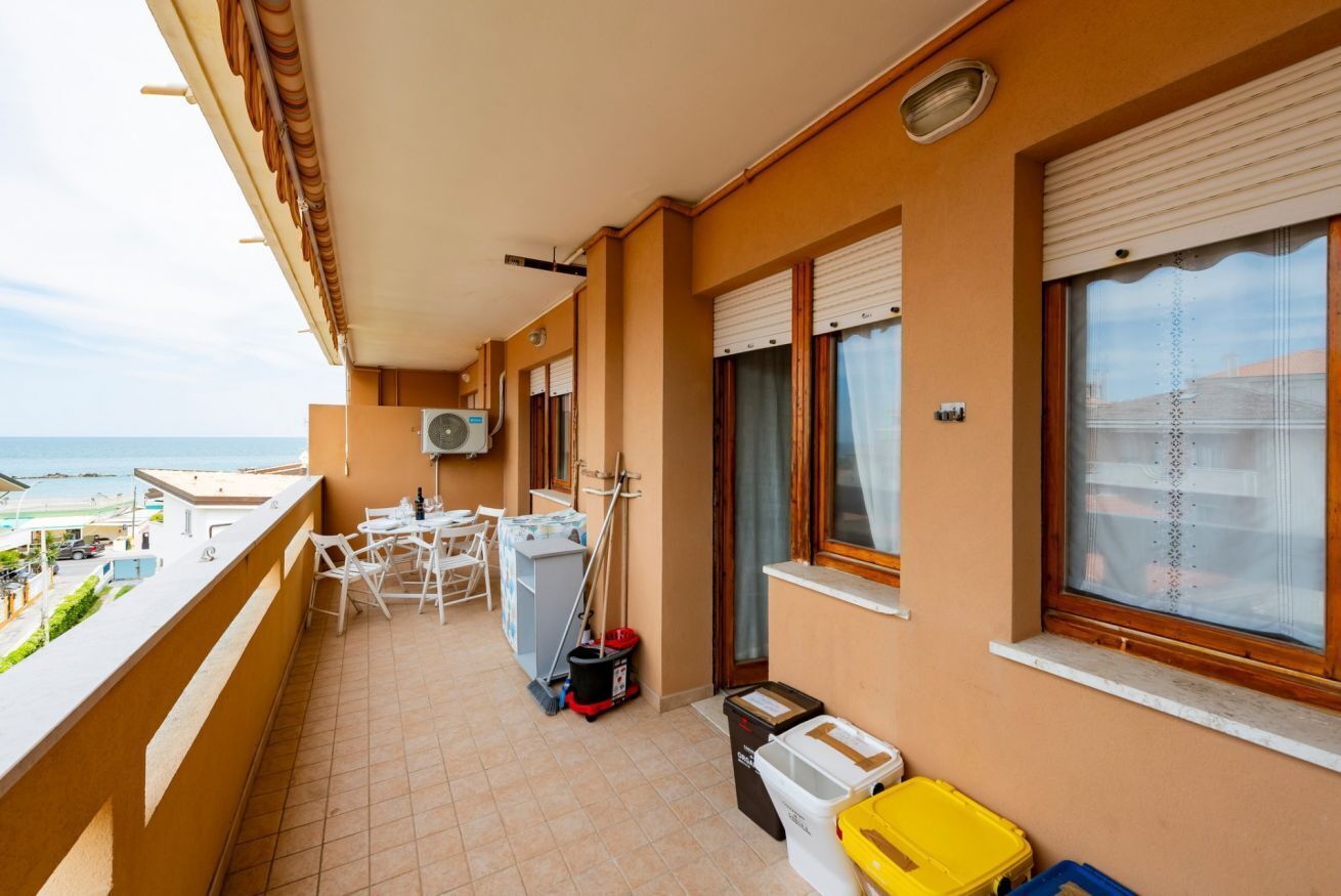 A balcony with a view of the sea, a small table with chairs, a window with blinds, and a trash bin.