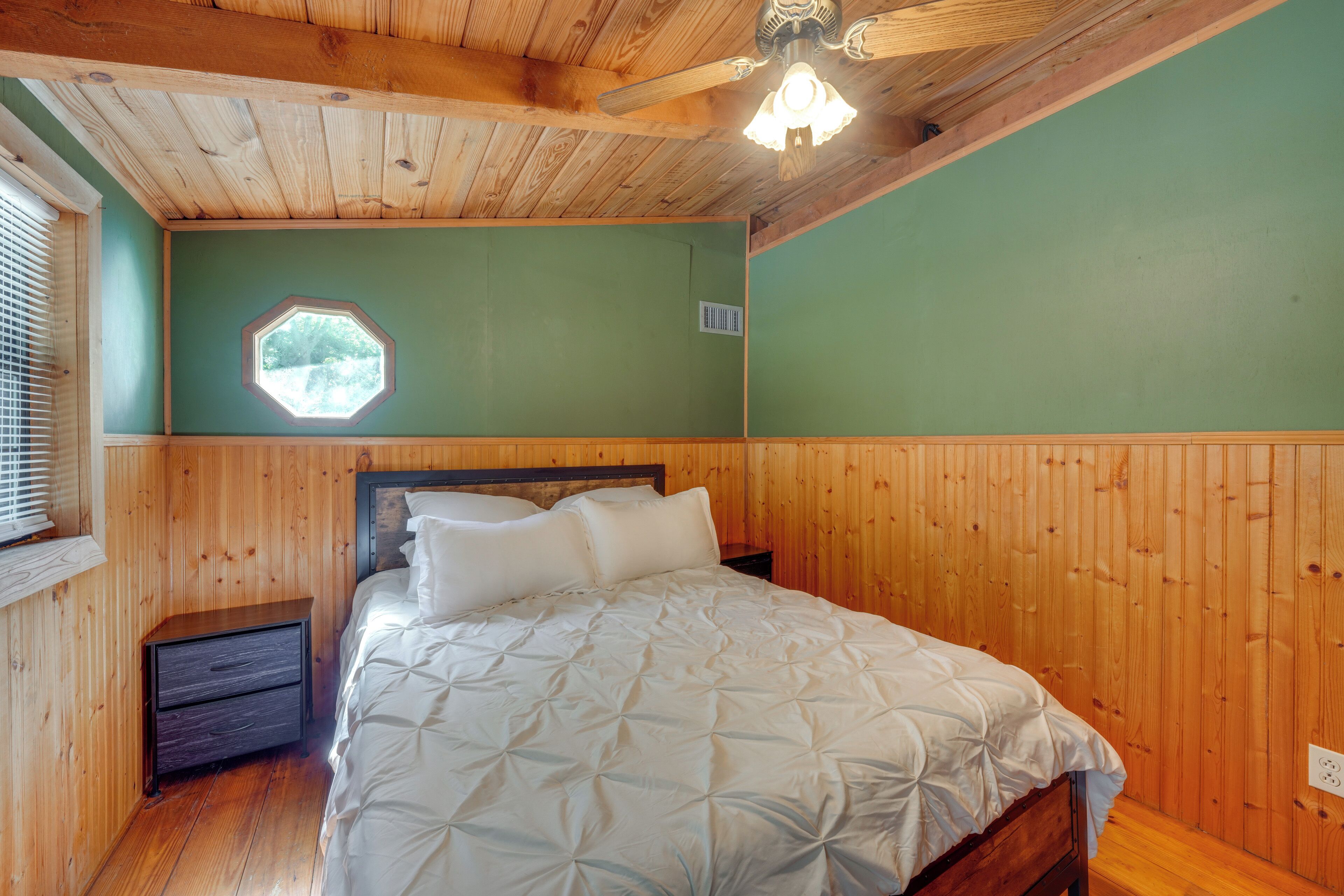 Un dormitorio con paneles de madera, una cama, una mesita de noche, una ventana con persianas y un ventilador de techo.