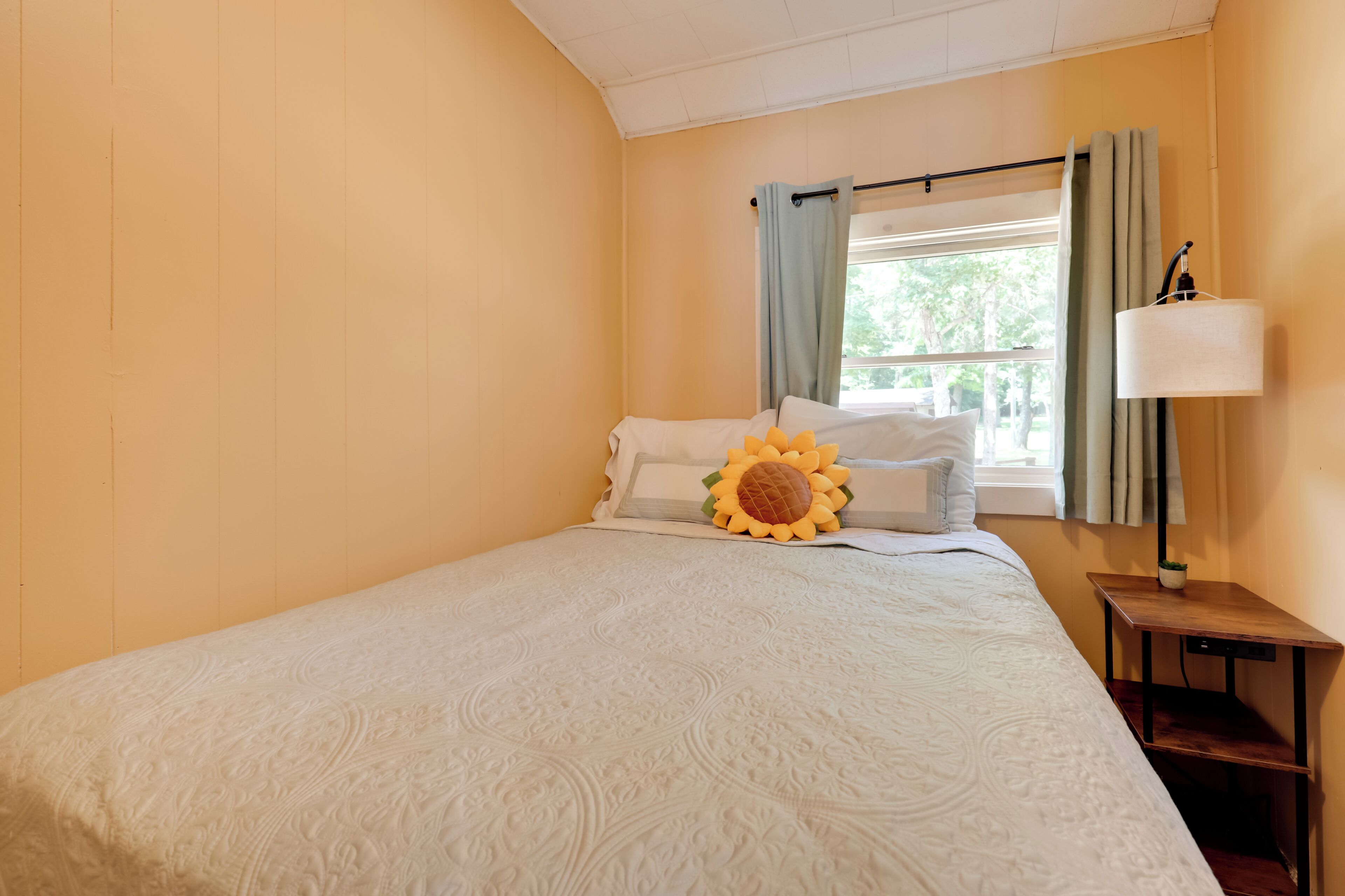 Una cama bien hecha con un almohadón de girasol, una lámpara de noche y una ventana con cortinas.