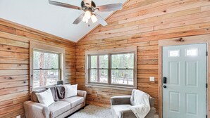Ein Blockhaus-Interieur mit Deckenventilator, zwei Fenstern, einer Tür, einer Couch und einem Sessel.