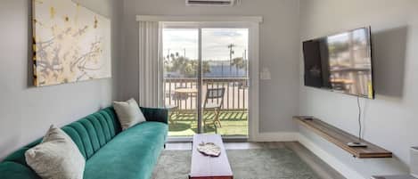 Un salon avec un canapé de couleur turquoise, une table basse, un téléviseur à écran plat et une grande fenêtre donnant sur l’extérieur.