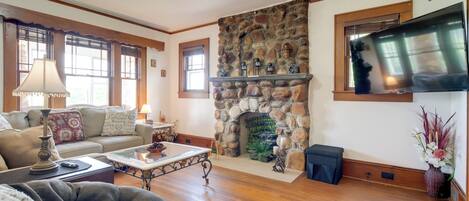 Una sala de estar acogedora con chimenea de piedra, pisos de madera, un sofá y un televisor montado en la pared.