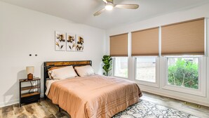 A bedroom with a bed, a nightstand, a ceiling fan, and large windows with blinds.