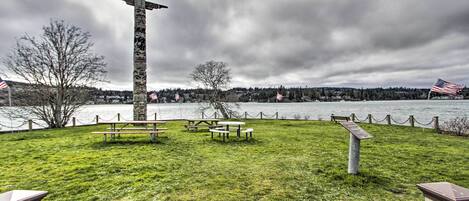 A park with picnic tables and a tall cross structure, overlooking a body of water and a forested area.