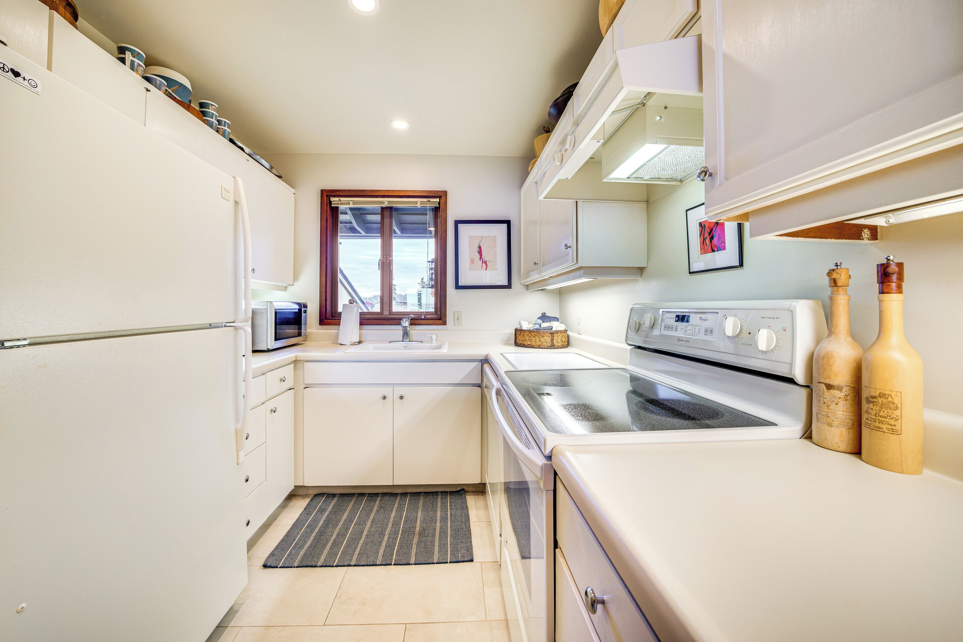 A compact kitchen with white cabinets, a microwave, a stove, and a sink. There are two wooden bottles on the counter and a window with a view of the city.