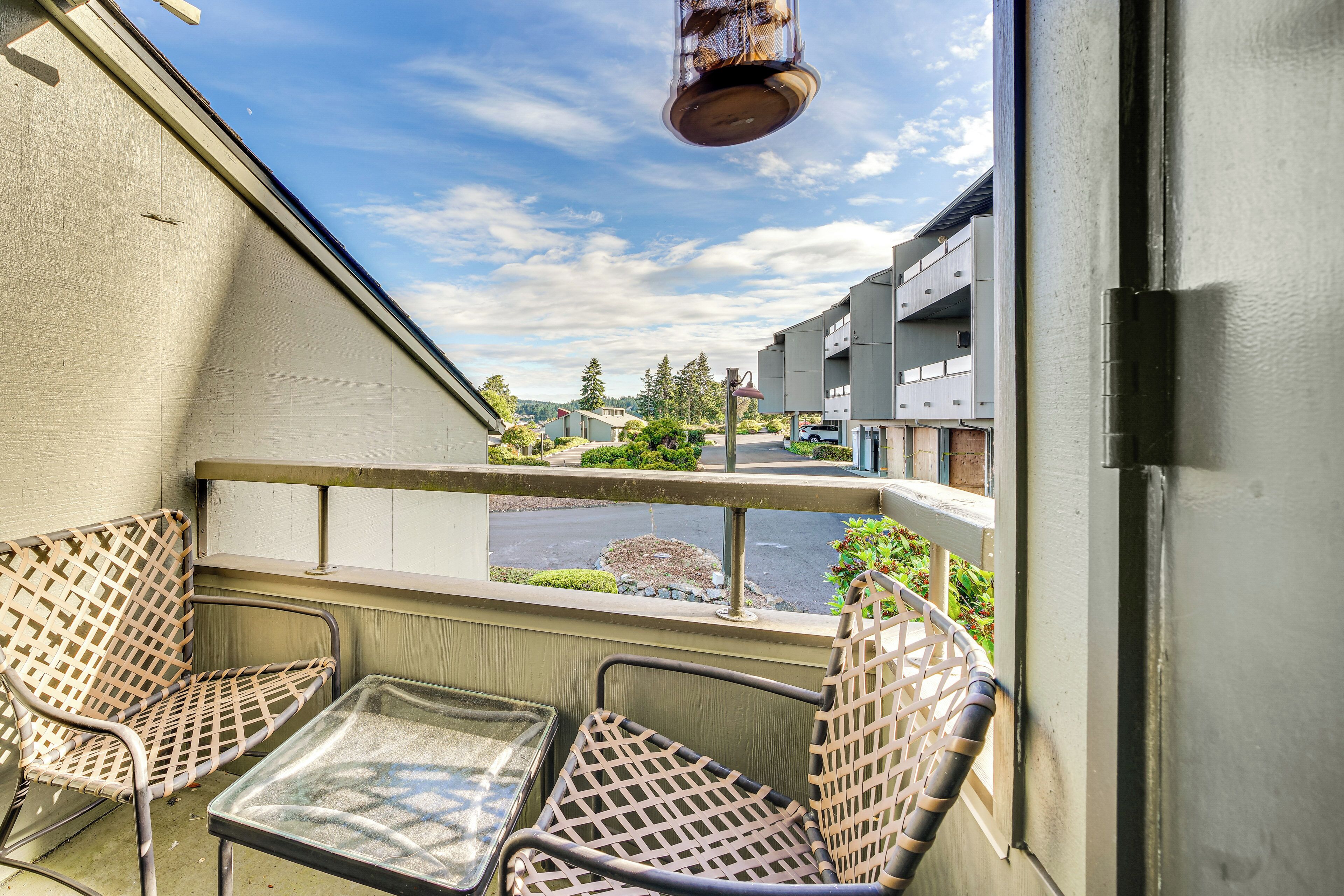 Un balcon avec deux fauteuils en osier et une petite table, donnant sur une rue bordée d’immeubles et d’espaces verts.