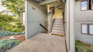 Una casa con escalera que lleva a un segundo piso, un pequeño porche y un jardín con arbustos y un árbol.