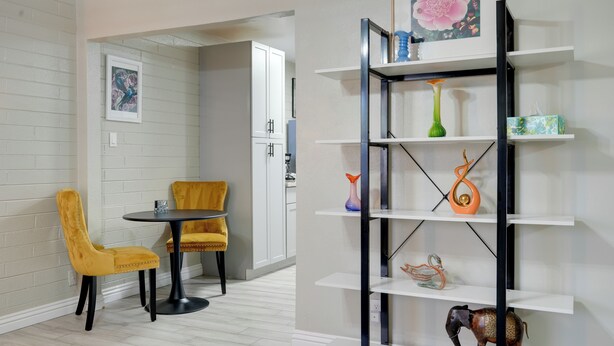 Een eethoek met een ronde tafel en twee gele stoelen, een wit kastje en een zwart metalen rek met decoratieve objecten.