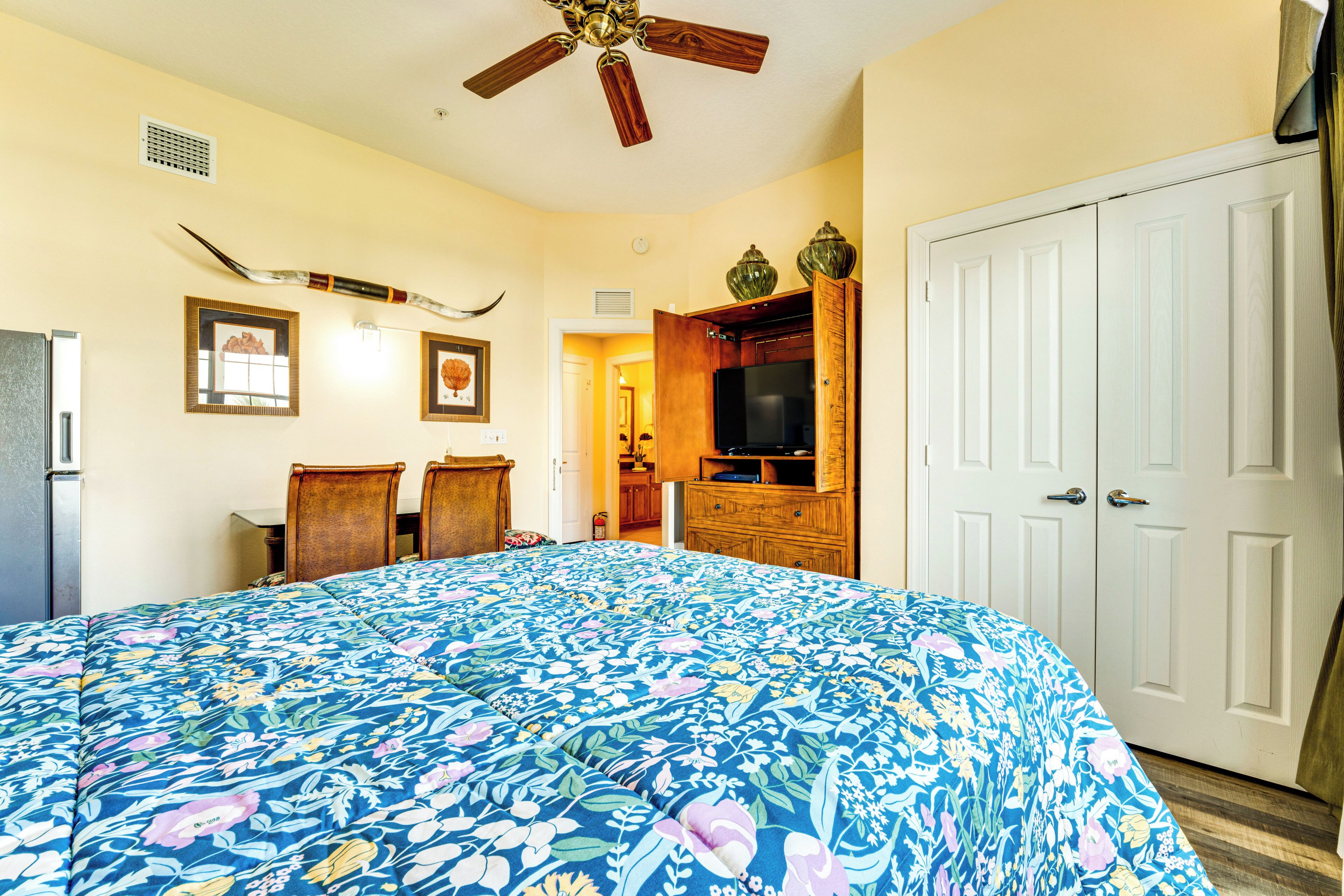 A bedroom with a large bed, a ceiling fan, a TV, wooden furniture, and a white door.
