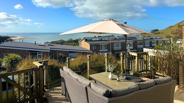 A seaside view with a patio area featuring wicker furniture and a large umbrella, overlooking a beach and residential buildings.