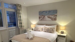 A bedroom with a large bed, two bedside lamps, a window with curtains, and a painting on the wall.