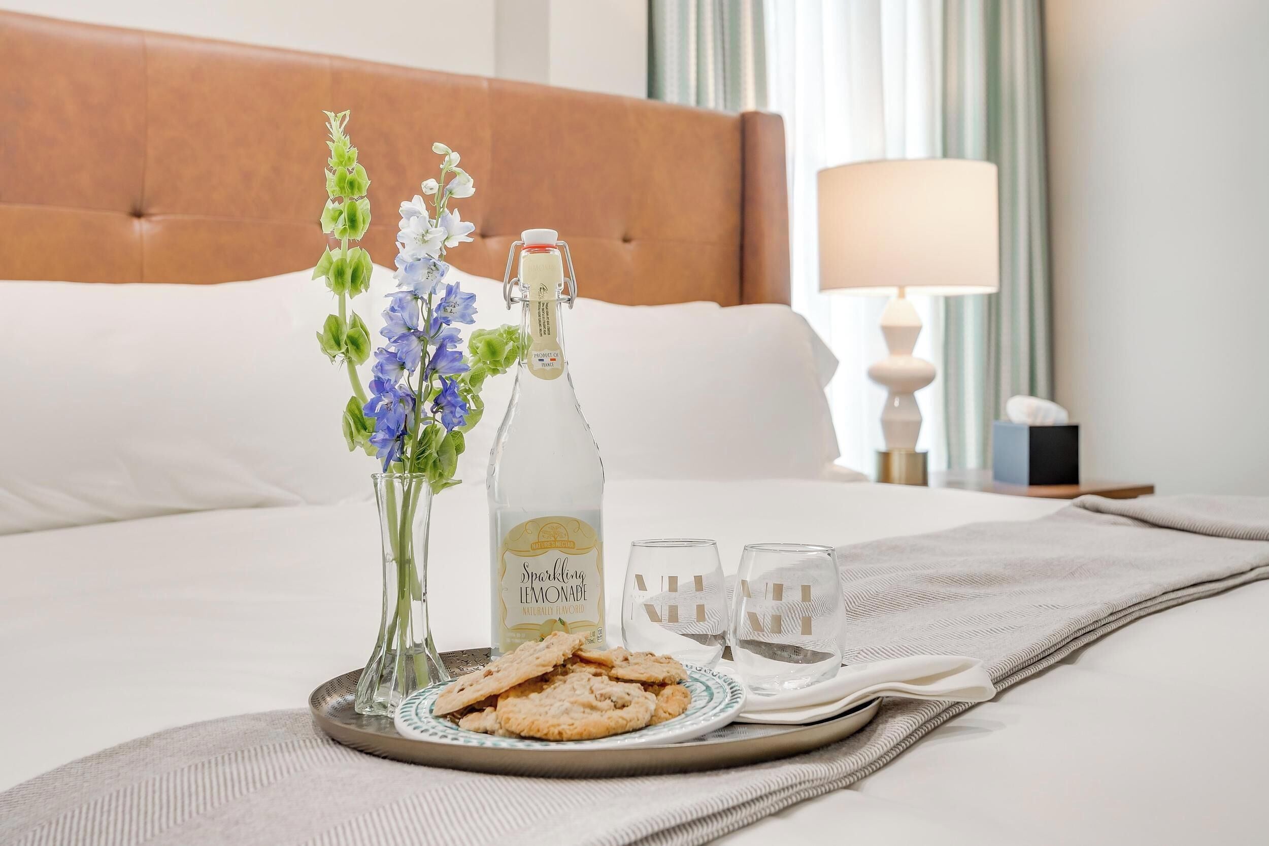 A neatly made bed with a brown headboard, a tray with snacks and a bottle of Sparkling Lemonade, and a vase with flowers.
