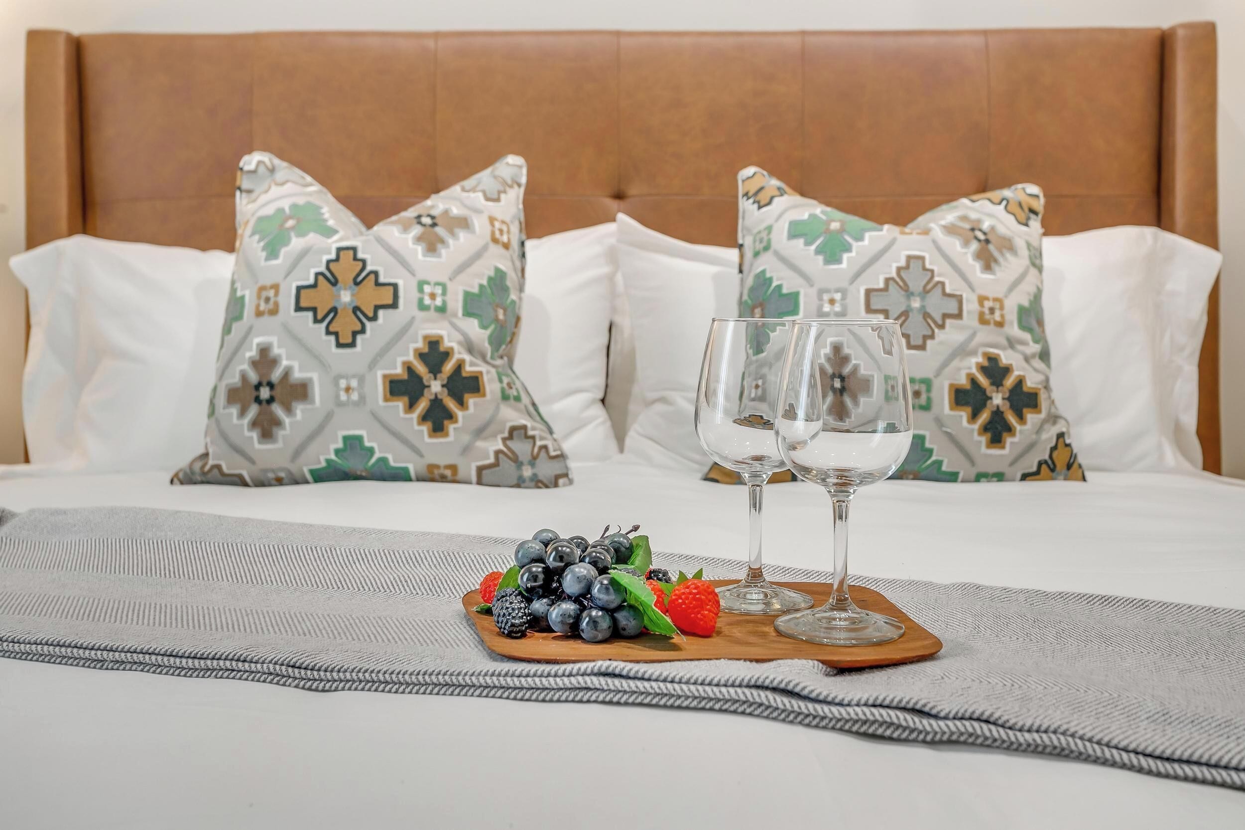 A neatly made bed with patterned pillows, a fruit platter, and two wine glasses on a tray.