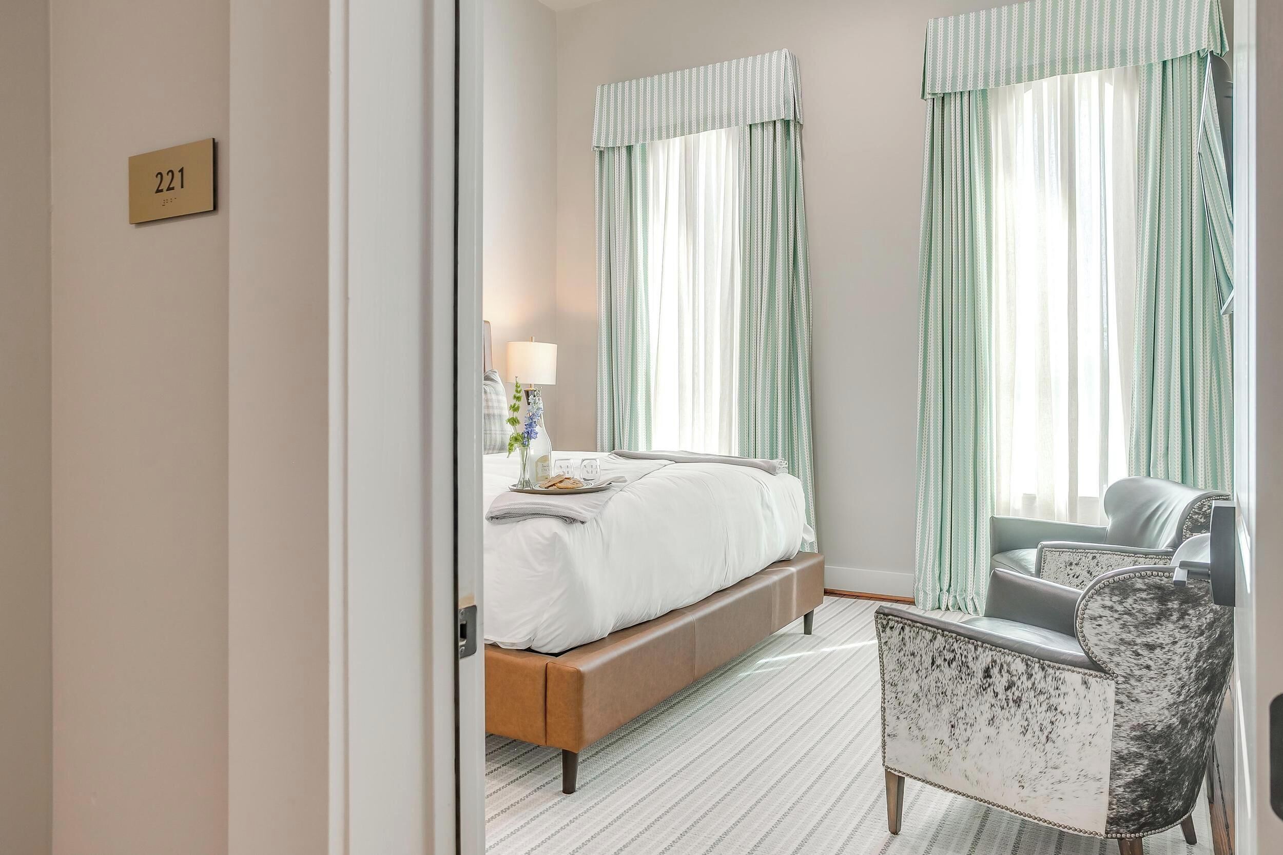 Habitación de hotel con cama, una silla, mesita de noche con lámpara y cortinas.