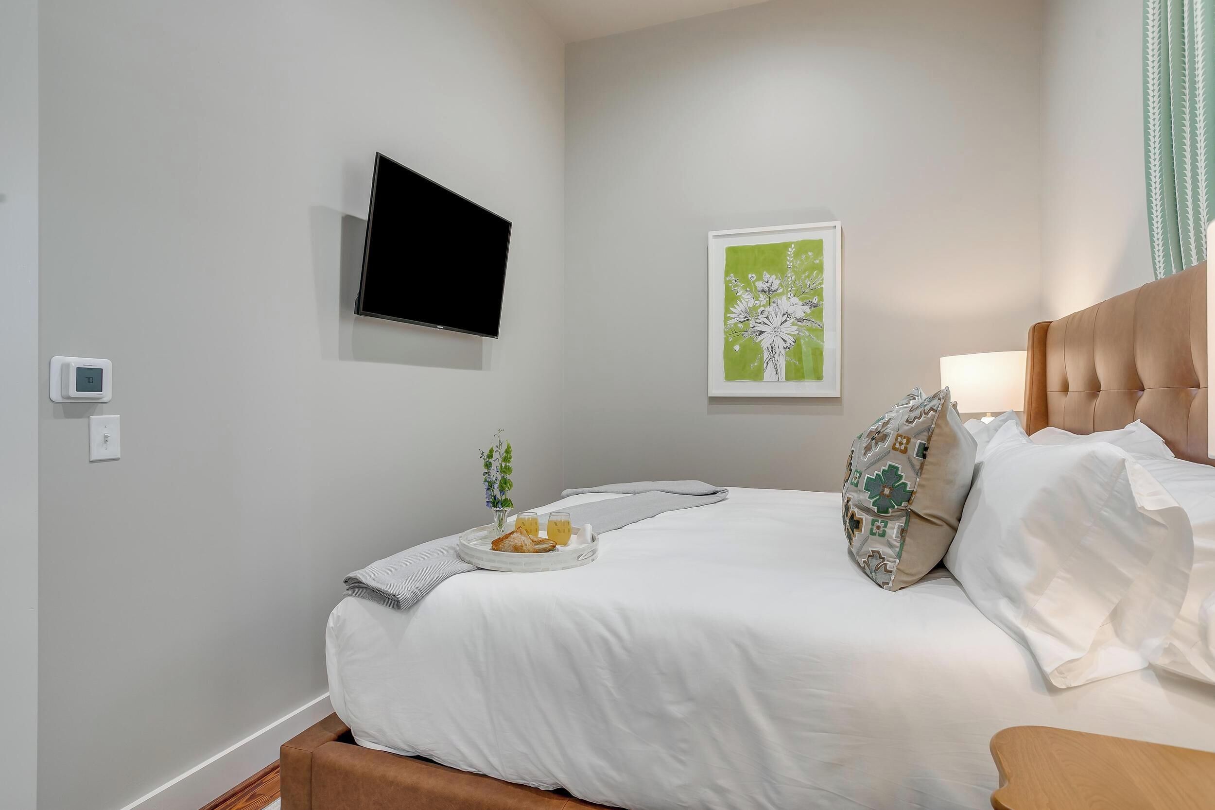 A neatly made bed with white linens, a bedside table with a lamp, a wall-mounted TV, and a framed floral artwork.