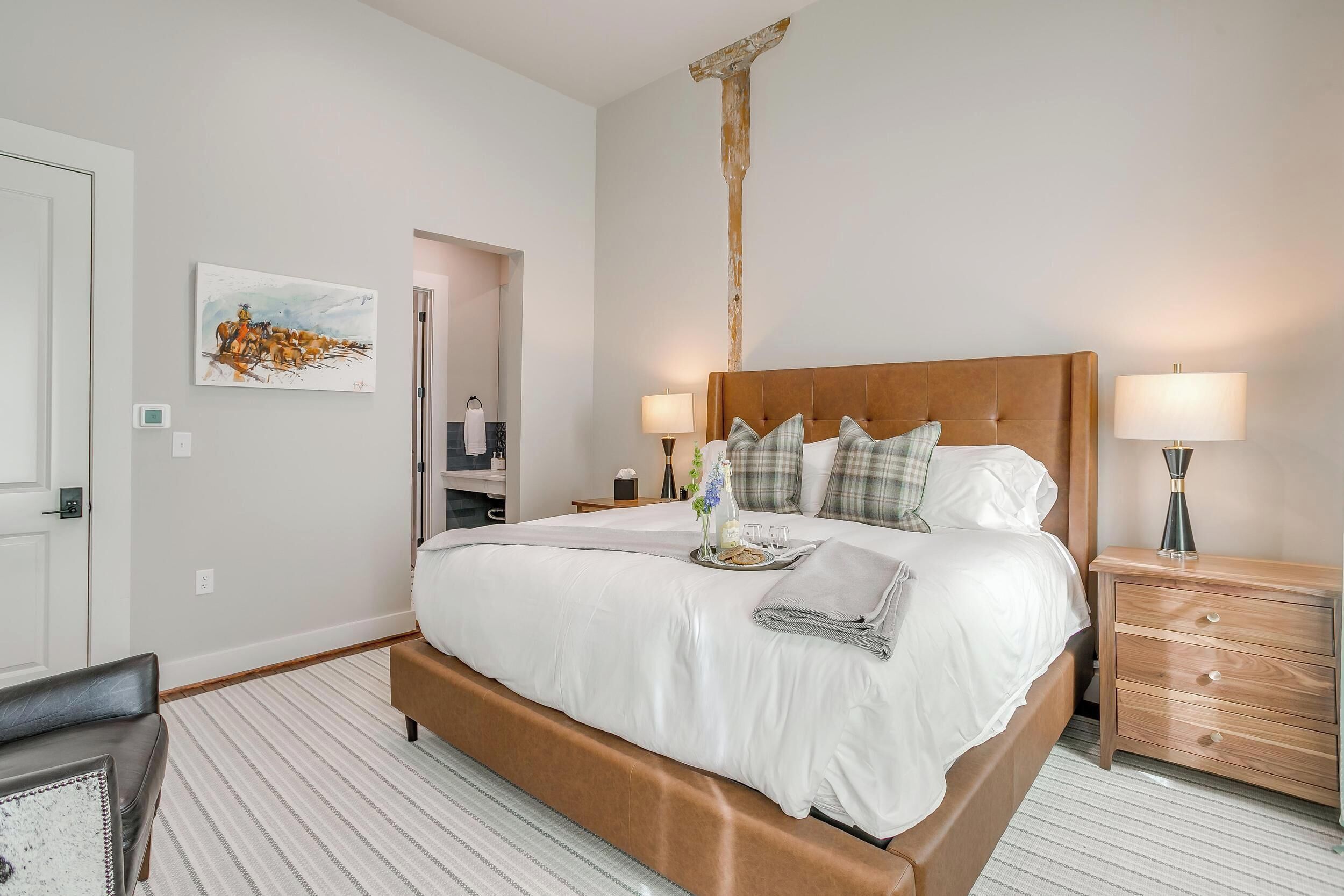 A neatly arranged bedroom with a large bed, bedside tables, a wooden nightstand, a painting on the wall, and a door leading to another room.