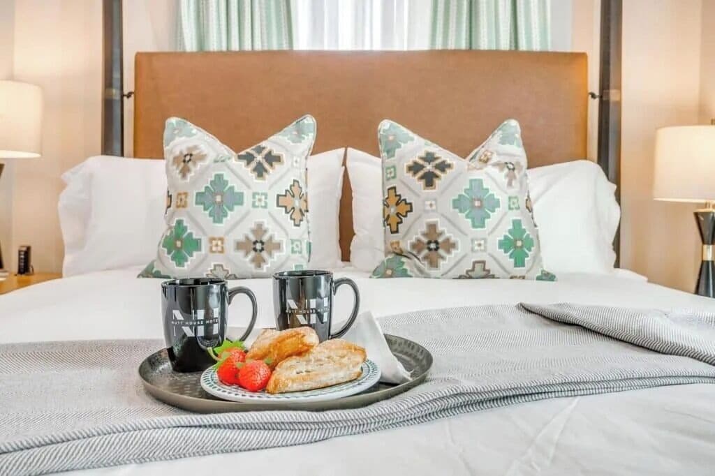 A neatly made bed with patterned pillows, a tray with a mug, a plate with toast and a strawberry, and a bedside lamp.