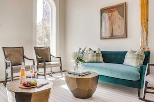 Un salón luminoso con un sofá verde, dos sillones, una mesa de centro con una botella y uvas, y un cuadro en la pared.