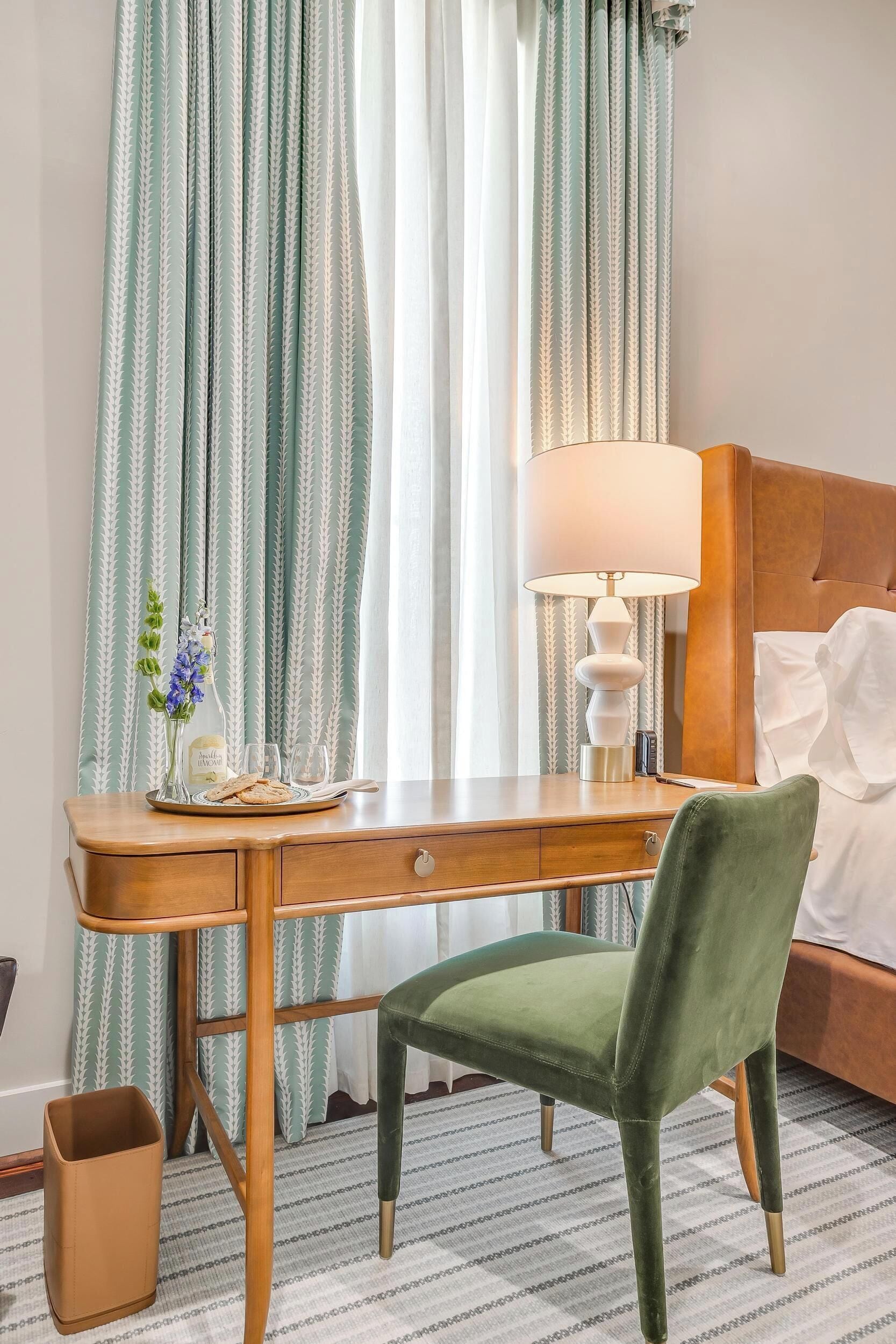 A hotel room with a wooden desk, a green chair, a bed with white linens, and a lamp.