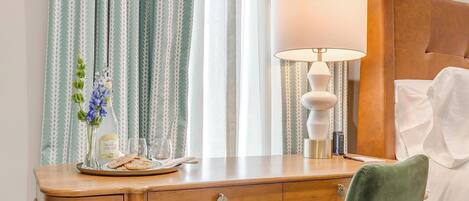A hotel room with a wooden desk, a green chair, a bed with white linens, and a lamp.