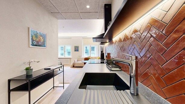A modern kitchen with a black countertop, stainless steel sink, and a herringbone-patterned backsplash. There is a dining area with a white sofa and a table, and a painting on the wall.