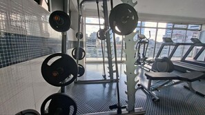 A gym with weightlifting equipment, a treadmill, and a view of buildings through large windows. - Omedad Hotel (Addis Ababa)