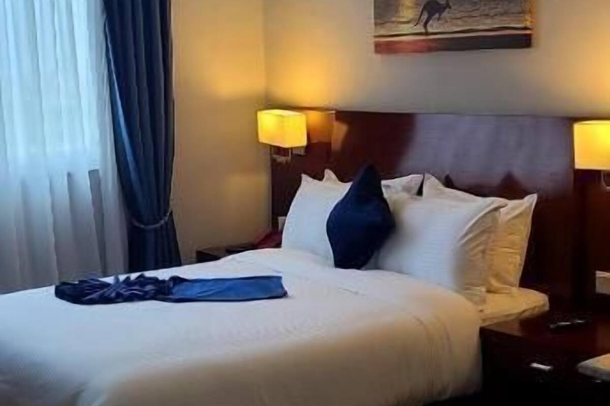 A hotel room with a bed, a nightstand, a lamp, a painting on the wall, and a window with curtains.