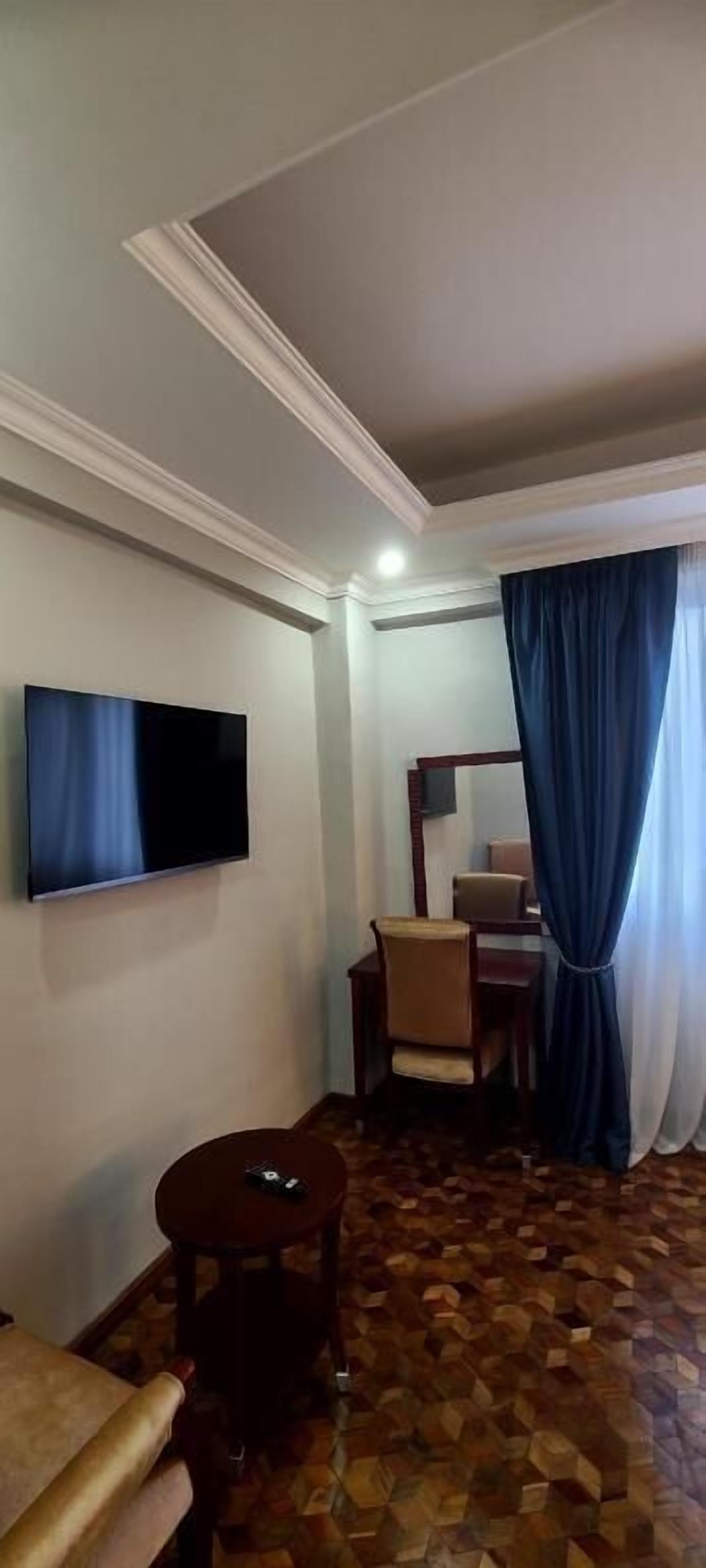 A hotel room with a flat-screen TV mounted on the wall, a round wooden table, a sofa, and a patterned floor.