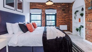 A modern bedroom with a brick wall, a large bed with white bedding, and a hanging light fixture. - Modern Flat in Central Leeds | Walk to City Centre (Leeds)