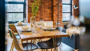 A dining table set with plates, glasses, and a vase of greenery in a room with brick walls and large windows. - Modern Flat in Central Leeds | Walk to City Centre (Leeds)