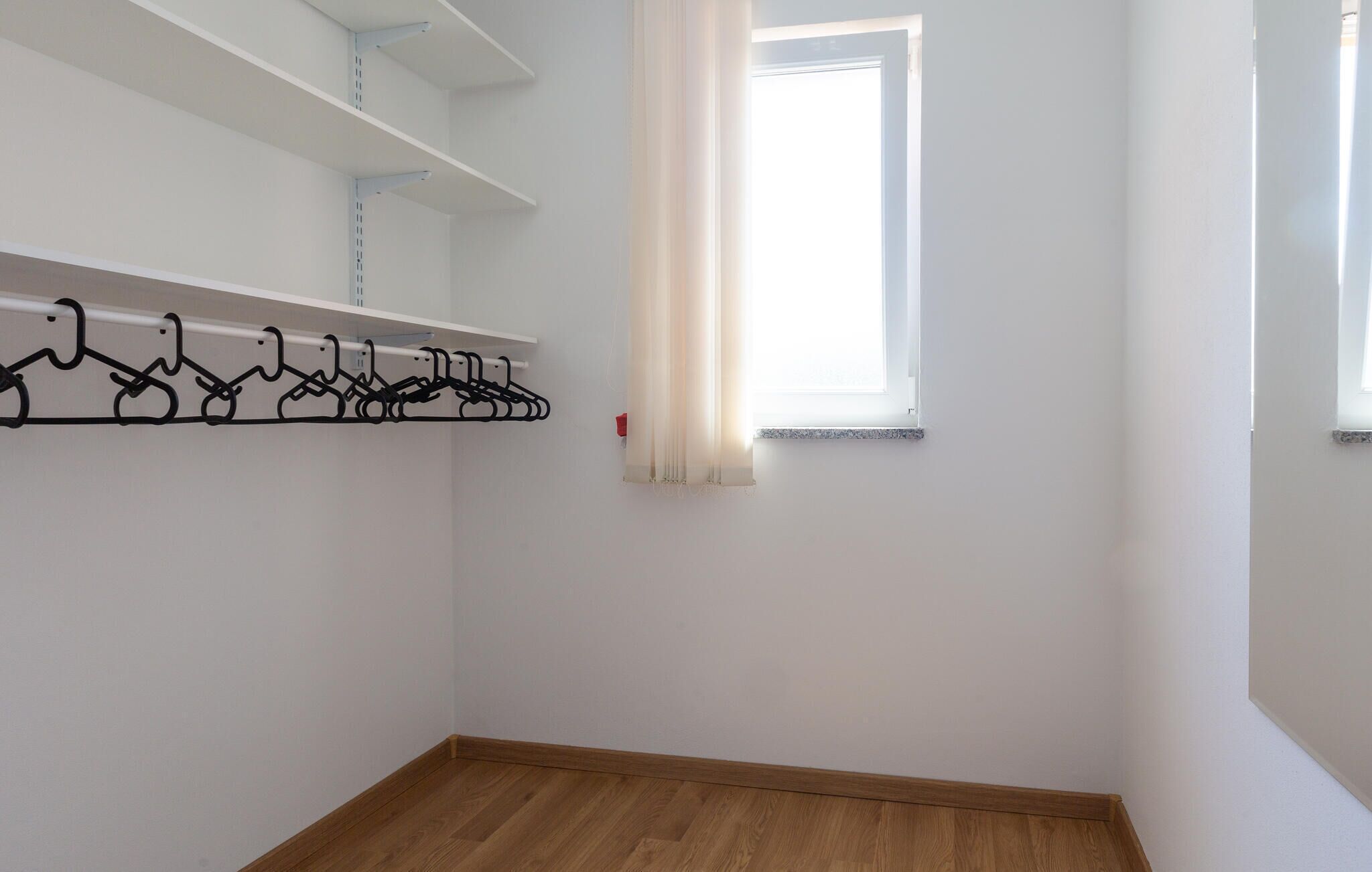 Une pièce avec un plancher en bois, des murs blancs, une fenêtre avec des rideaux blancs et un porte-manteau noir.