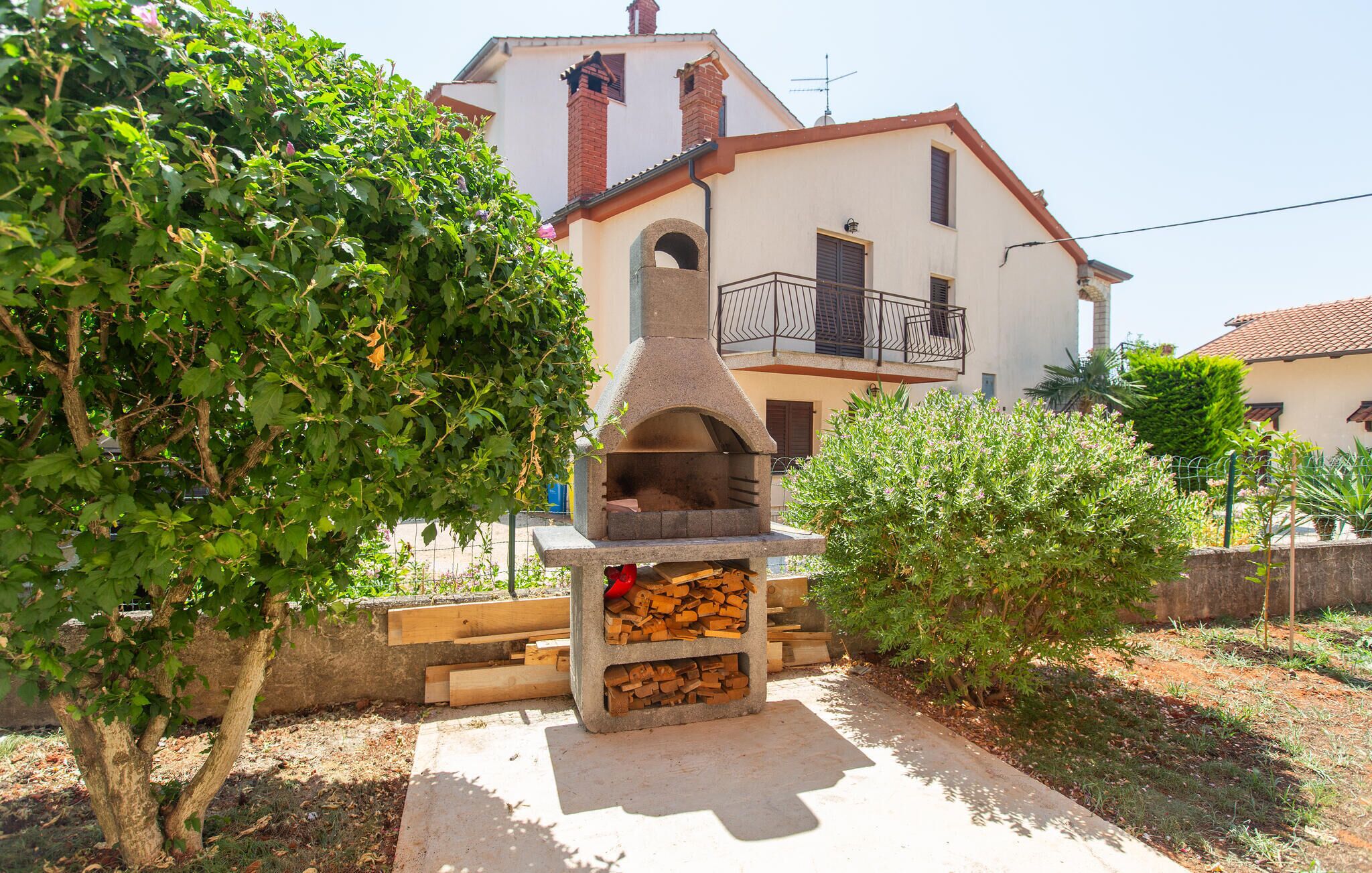 Un horno de piedra tradicional al aire libre con leña apilada debajo, rodeado de abundante vegetación y un edificio de dos plantas con balcón al fondo.