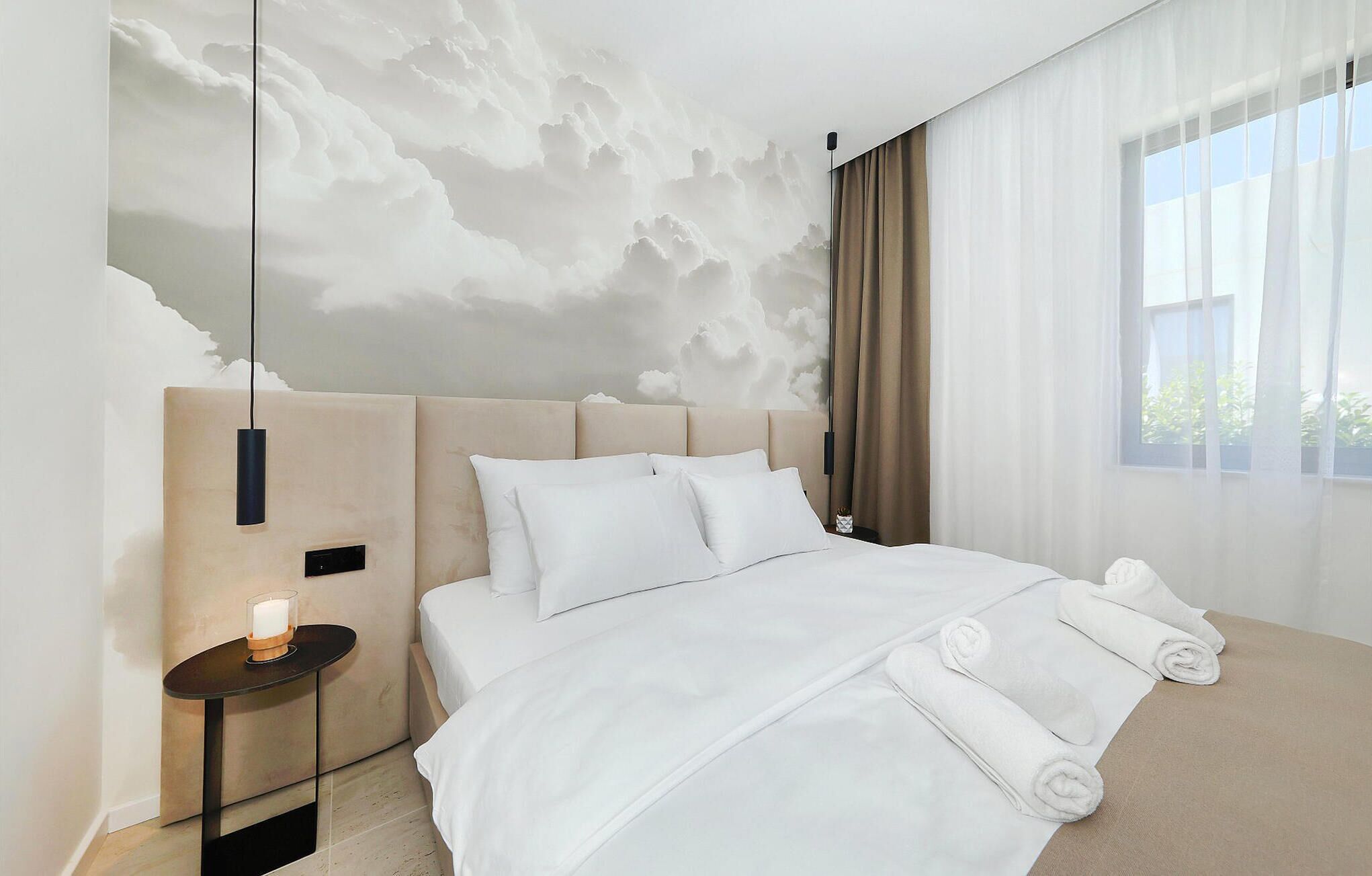 A modern hotel room with a large bed, a bedside table with a candle, and a view of the sky through sheer curtains.