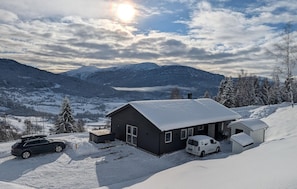 A snow-covered cabin with a car parked in front, surrounded by mountains and a clear sky.