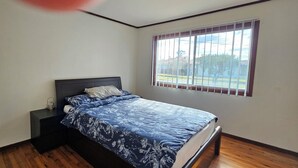 Un dormitorio con cama, mesita de noche, ventana con persianas y vistas al exterior.