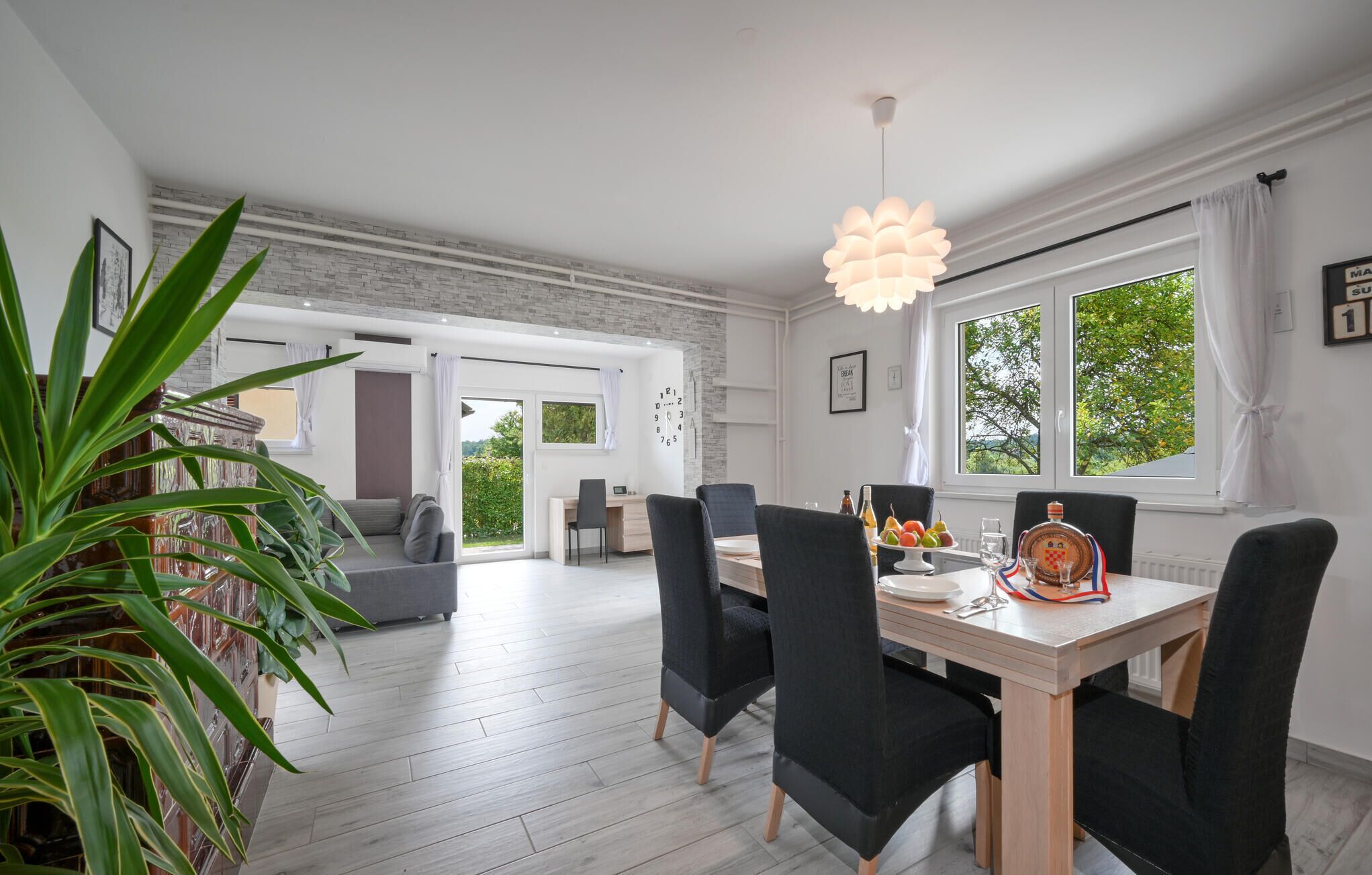 Ein zeitgemäßer Essbereich mit einem Holztisch, schwarzen Stühlen und einer Pendelleuchte. Ein großes Fenster mit Vorhängen bietet Blick auf Grünflächen, und eine Treppe führt in einen anderen Raum.