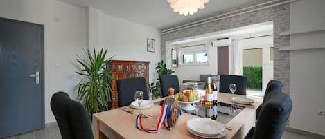 Un coin repas avec une table en bois, dressée pour quatre personnes, entourée de chaises. On y trouve une grande plante sur la gauche, un panneau décoratif au mur et un luminaire suspendu moderne au-dessus de la table.
