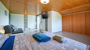 Un dormitorio con techo de madera, una cama con ropa de cama estampada, un sofá gris, una mesita y un armario.