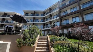 A multi-story residential building with balconies, a staircase, and a garden area with shrubs and a tree.