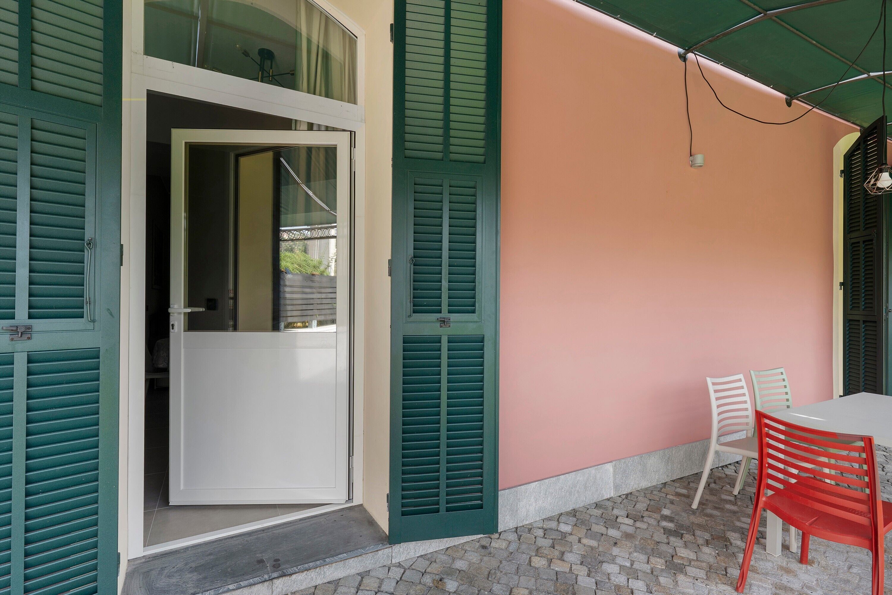 Una habitación con puerta blanca, contraventanas verdes, pared rosa y una mesa con dos sillas.