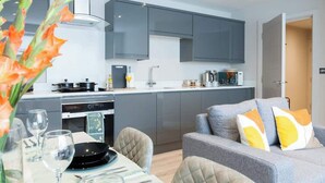 Une cuisine moderne avec des meubles gris, un coin repas avec une table dressée pour deux personnes, et un canapé gris agrémenté de coussins jaunes.