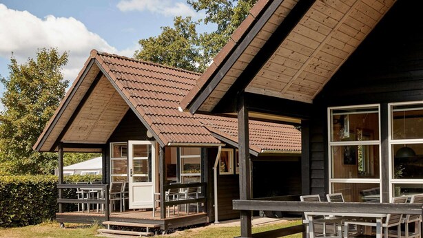 A wooden cabin with a sloped roof, a porch with seating, and large windows. - 6 Person Holiday Park Home in Bryrup (Bryrup)