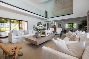 A spacious living room with a high ceiling, a large white sofa, a coffee table with decorative items, and a staircase leading to an upper level.