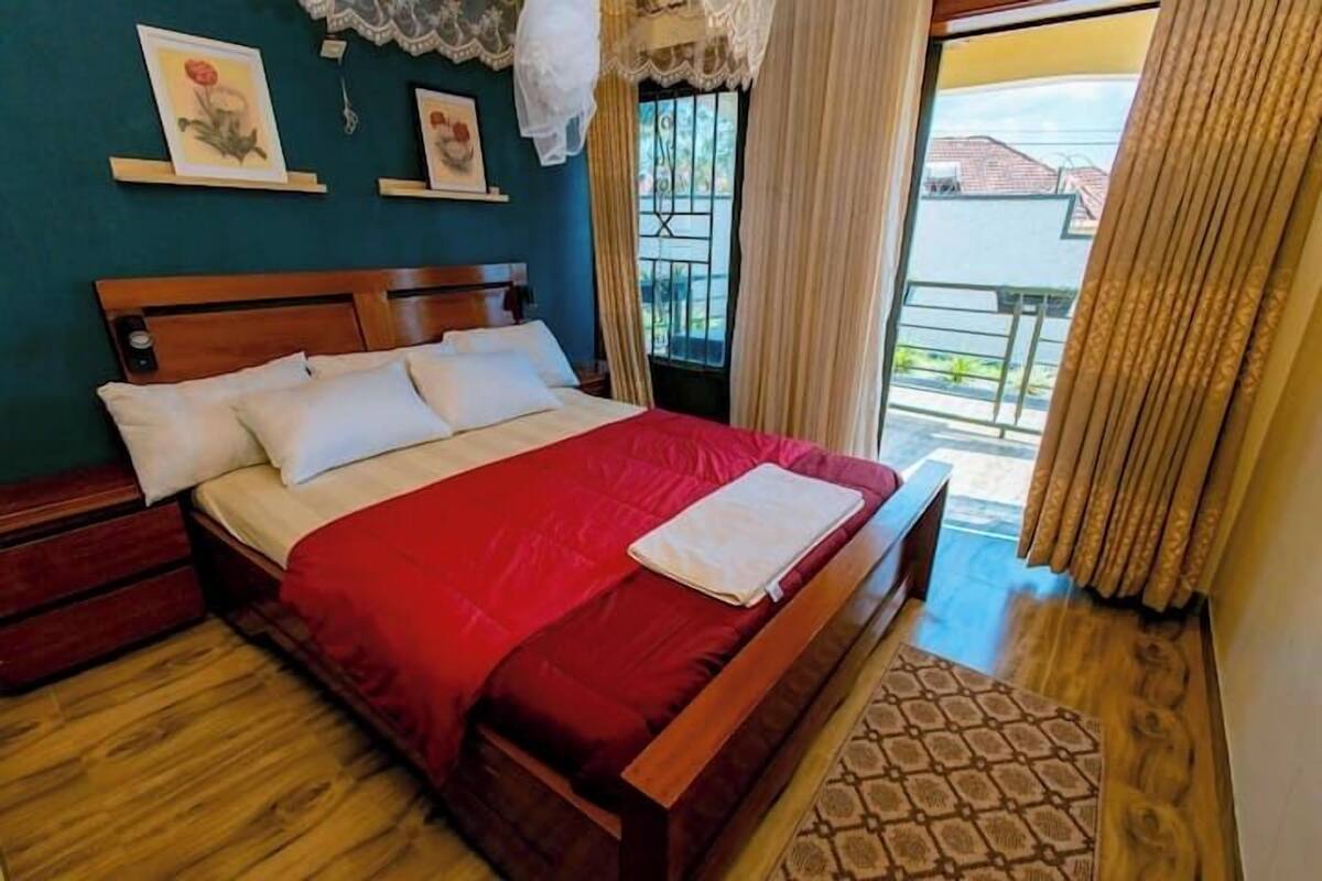 A neatly made bed with a red bedspread and white pillows, a wooden headboard, and a view of a balcony with a railing.
