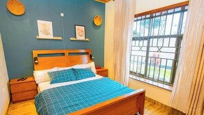 A bedroom with a wooden bed, a nightstand, a bedside table, a window with a view, and wall decorations. - Homescapes (Kigali)