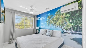 Een slaapkamer met een bed, een plafondventilator, een airconditioning en een raam met gordijnen, plus een wanddecoratie van een tropisch strand.