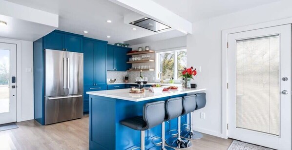 Una cocina moderna con armarios azules, un refrigerador de acero inoxidable, una isla central con taburetes y un ventanal que proporciona luz natural.