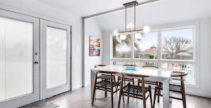 Un comedor con mesa blanca y cuatro sillas, ventanas grandes con persianas y una lámpara colgante.